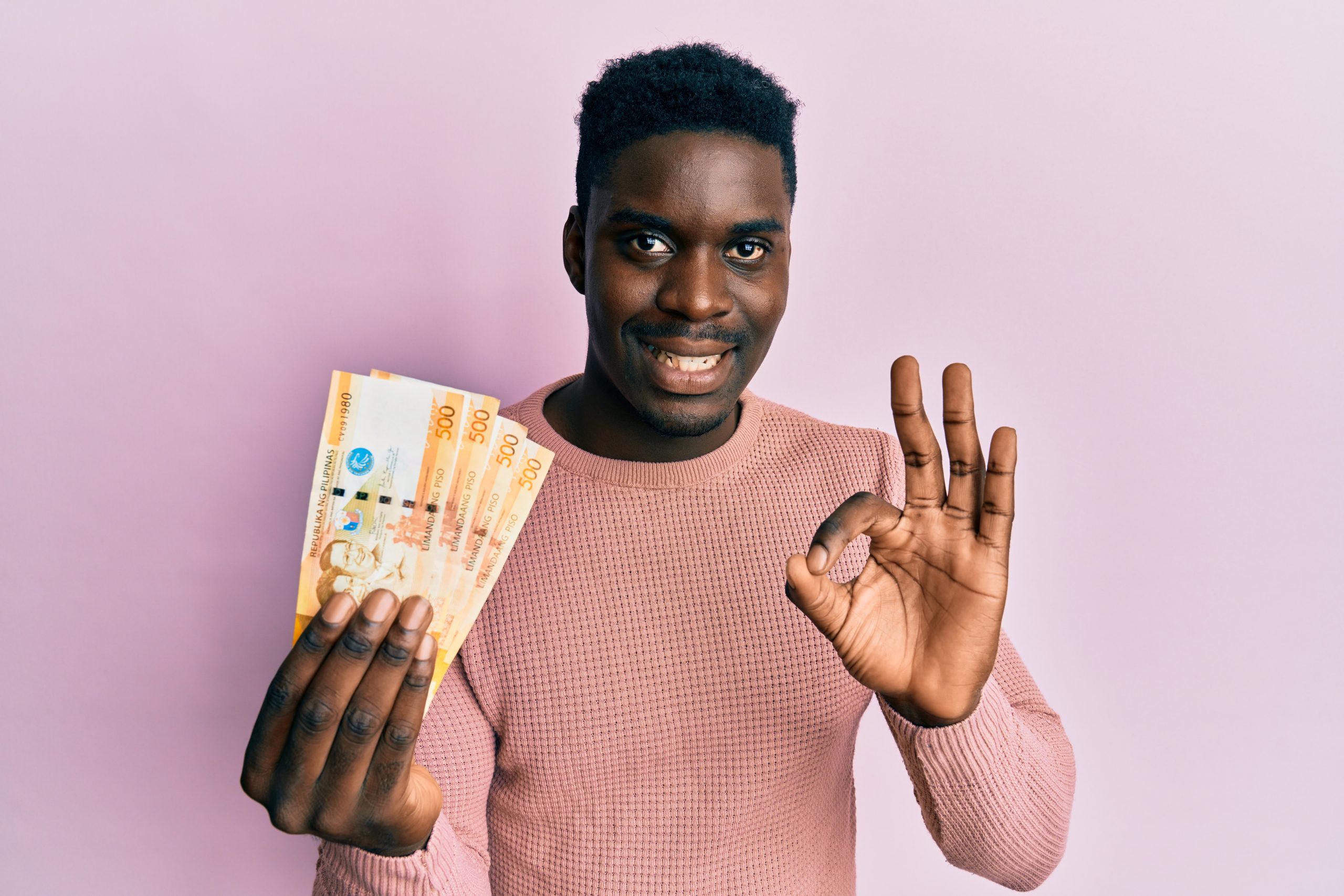 Handsome black man holding 500 philippine peso banknotes doing ok sign with fingers, smiling friendly gesturing excellent symbol